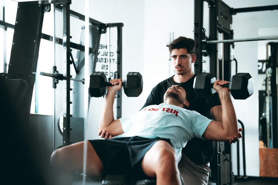 Treinador do ShapeUp a apoiar um aluno no supino com halteres